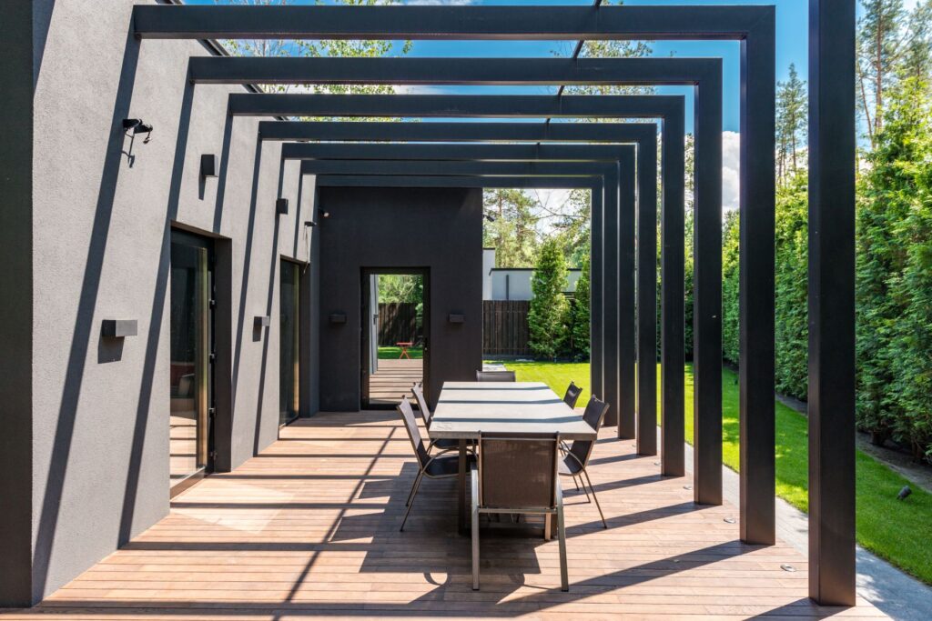 Åpen pergola ved husvegg med spisebord og tett grønn hekk