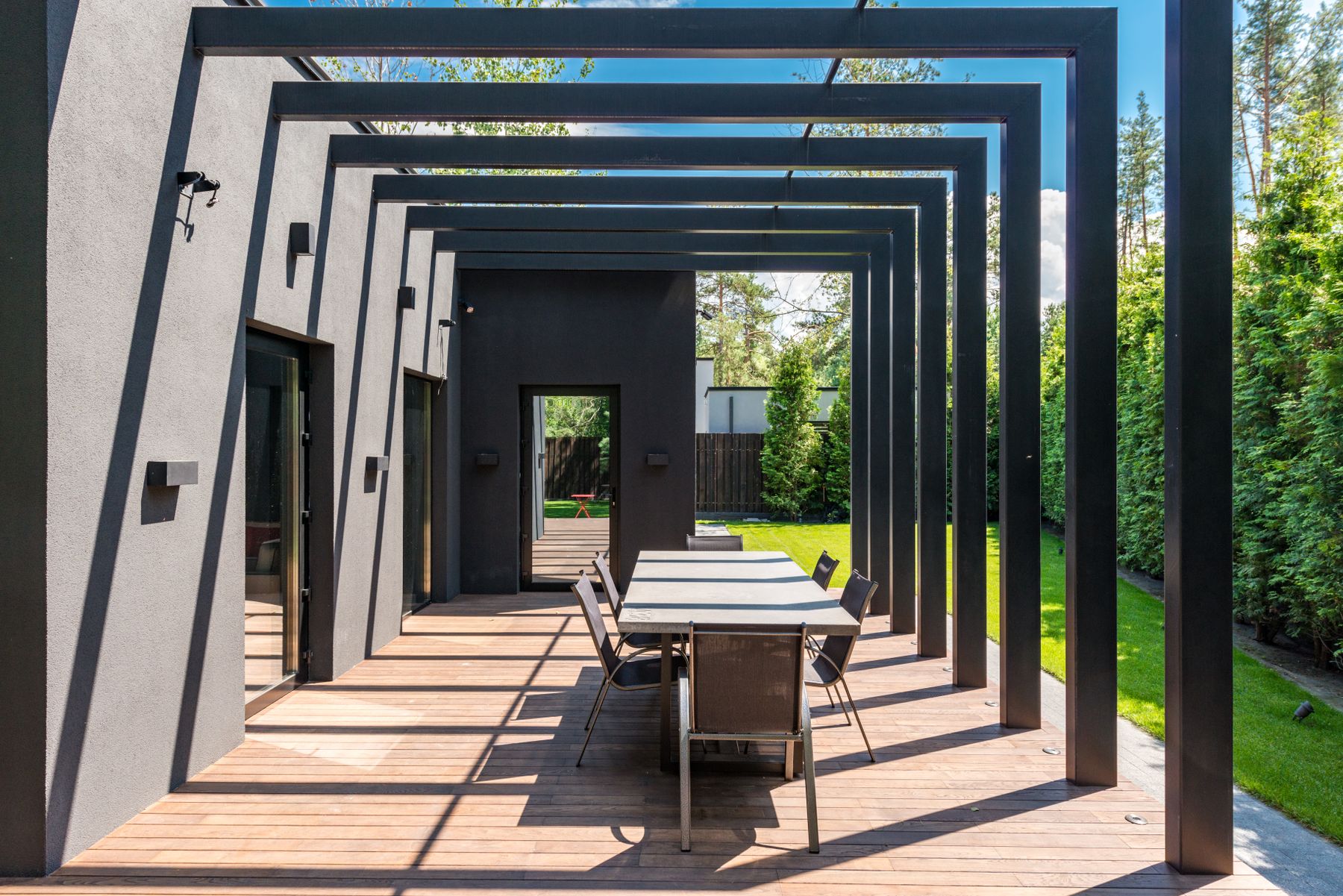 Åpen pergola ved husvegg med spisebord og tett grønn hekk