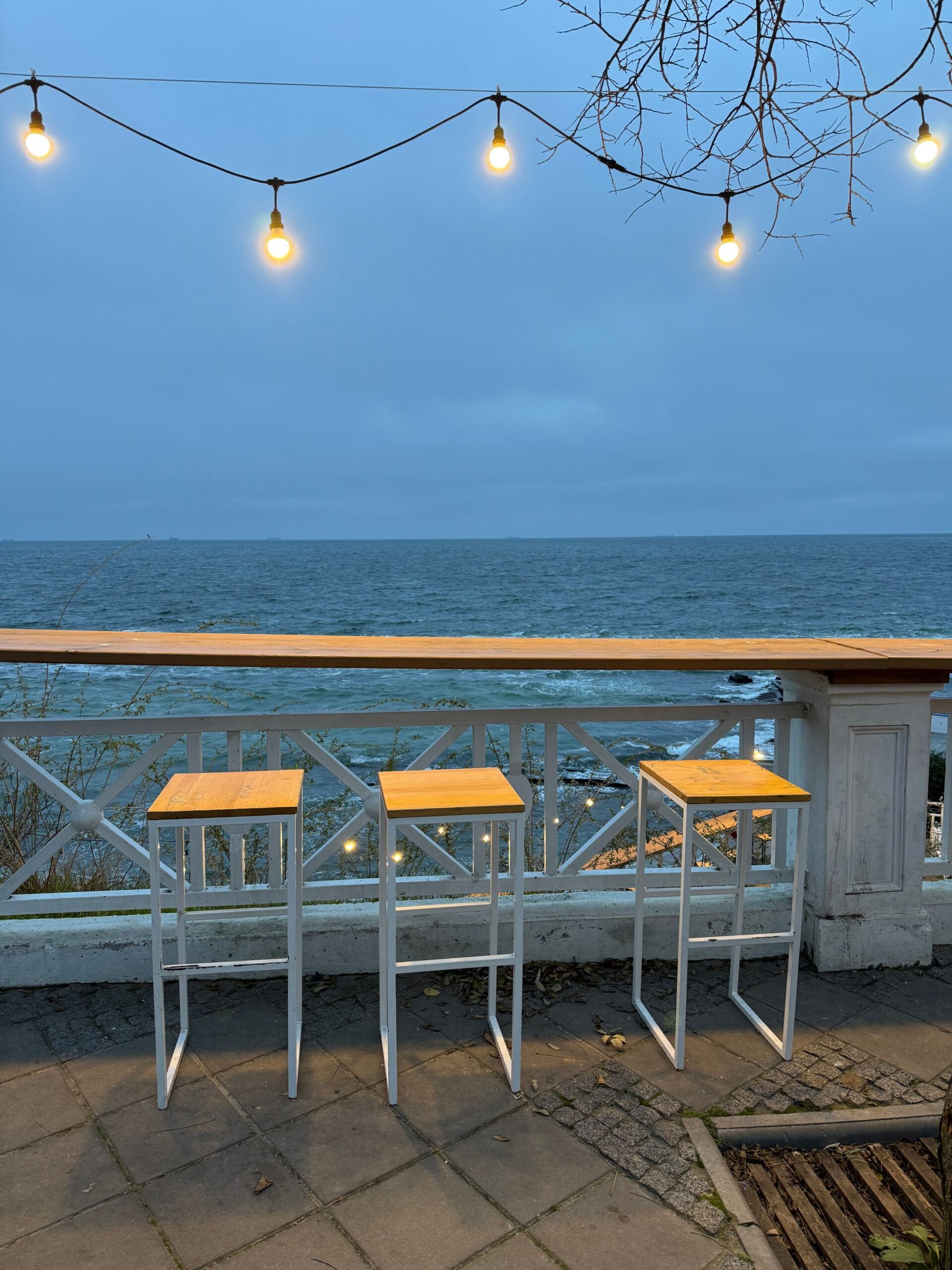 Lyslenke over en åpen terrasse med høye stoler og utsikt mot havet i skumringen